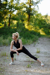 Pretty caucasian young blonde girl in black sportswear doing fitness exrecise at sunset in summer park. Warming up outdoor.