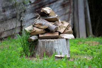 Pile of firewood stacked on top of large tree stump surrounded with uncut grass and barn in background on warm sunny spring day