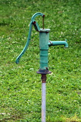 Vintage old rusted green wrought iron hand water pump mounted on single metal pipe in local garden surrounded with uncut grass on warm spring day