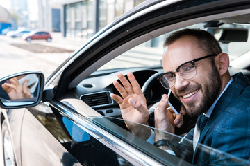 selective focus of happy bearded businessman in glasses talking on smartphone while waving hand in car