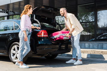 low angle view of happy man putting pink luggage in car trunk near woman