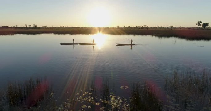 Aerial view of three polers silhouetted against the setting sun rowing their Mokoros on the waterways of the Okavango Delta