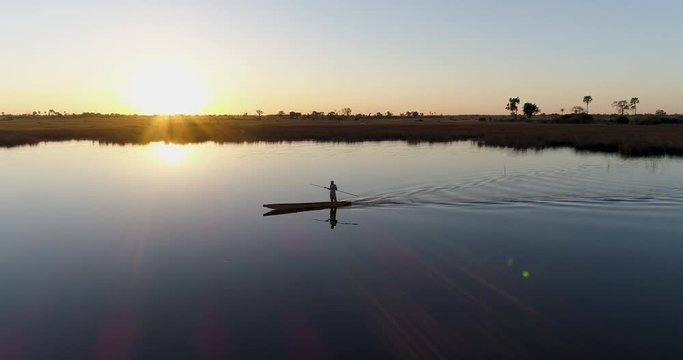 Aerial close-up panning view of one poler silhouetted against the setting sun rowing a Mokoro on the waterways of the Okavango Delta