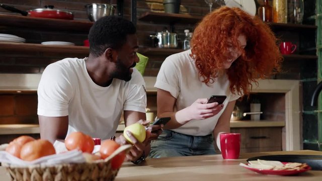 Young african American man eat apple with curly red woman use their smart phone couple happy smile talk at home at kitchen girlfriend internet browsing technology together date shopping slow motion