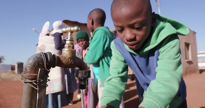Water crisis.Young african boys drinking water from a tap while woman line up to collect water in plastic containers due to severe drought in South Africa