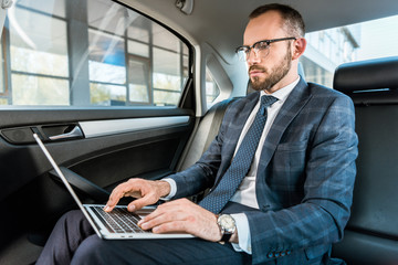 low angle view of handsome businessman in glasses typing on laptop in car