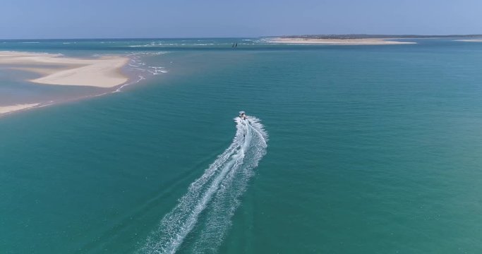 High aerial view of a pretty young girl wakeboarding pulled by a speedboat on an beautiful scenic estuary with dhow sailing boats in the background