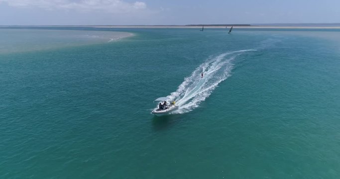 Aerial front view of a pretty young girl wakeboarding pulled by a speedboat on an beautiful scenic estuary with dhow sailing boats in the background