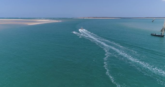 Aerial back view of a pretty young girl wakeboarding pulled by a speedboat on an beautiful scenic estuary with dhow sailing boats in the background
