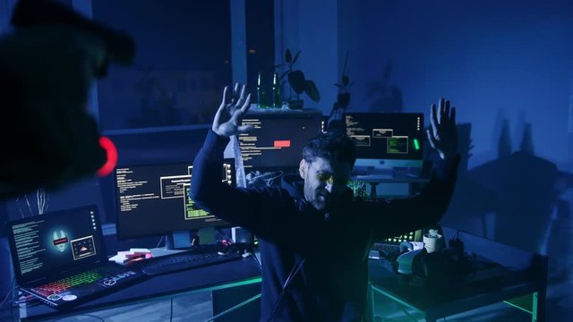 View of arrested internet hacker raising hands surrendering to the police. Computer hacker hideout in dark room. Cyber crime and malware concept.