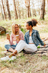 two multiethnic friends sitting on plaid blanket and holding paper cups of coffee