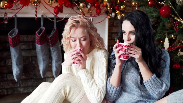 Beautiful brunette and women in warm sweater and socks are drinking hot chocolate with marshmallows near Christmas tree