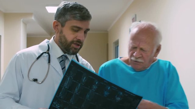Tilt up and down shot of doctor walking through hospital hallway with old patient with cane and explaining the results of X-ray, showing picture and talking about diagnosis