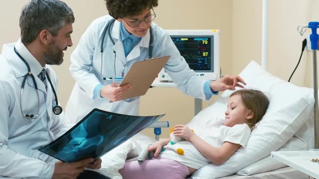 Male and female doctors standing with X-ray pictures near therapy bed of child, who holding soft toy and explaining her therapy