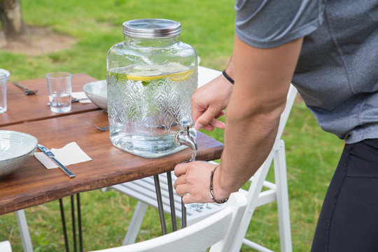 Closeup of man pouring lemonade from dispenser outdoors. Beverage dispenser on wooden table on green lawn with chairs. Summer drink concept.