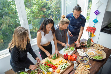 Naklejka premium Serious people cooking vegetables at kitchen table. Man and women standing at table with fresh vegetables and window with green view in background. Healthy cooking concept.