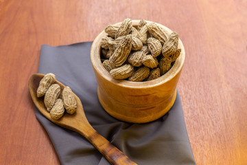 Peanut with peel and peel in a bowl next to a wooden cutlery and tablecloth