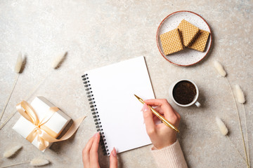 Modern minimal home workspace desk with human hand writing text message on blank paper notebook, waffles, dry flowers on concrete stone background. Flat lay style, top view fashion lifestyle blog hero