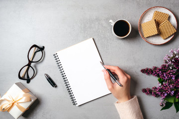 Top view hand of woman writing text message on blank paper notebook on stone table. Flat lay lilac flower, feminine stuff, coffee cup, glasses, gift box. Modern minimal home workspace desk concept.