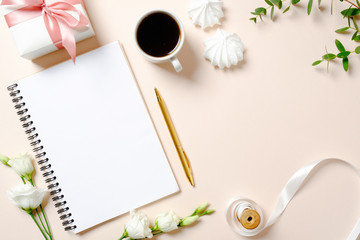 Flat lay home office desk. Female workspace with blank paper notebook, coffee cup, gift box, marshmallows and roses flowers on pink background. Top view feminine background with copy space for text.
