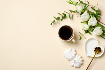 Flat lay morning coffee cup, marshmallow, white roses flowers and breakfast accessories on pastel beige color background. Top view women desk with feminine stuff. Beauty blogger banner concept.