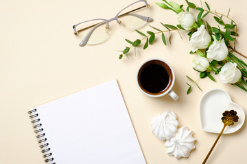 Flat lay feminine accessories, glasses, blank paper notepad, coffee cup, marshmallow and white roses flowers on pastel colors beige background. Top view beauty blogger desk with diary and female stuff