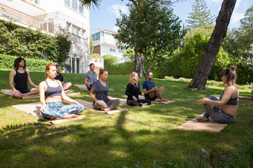 Team of people sitting in lotus pose on grass. Yoga trainer instructing trainee during outdoor workout. Practicing yoga concept