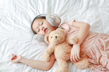Cute little Asian girl wearing winter ear muffs with teddy bear lying on the bed at home.
