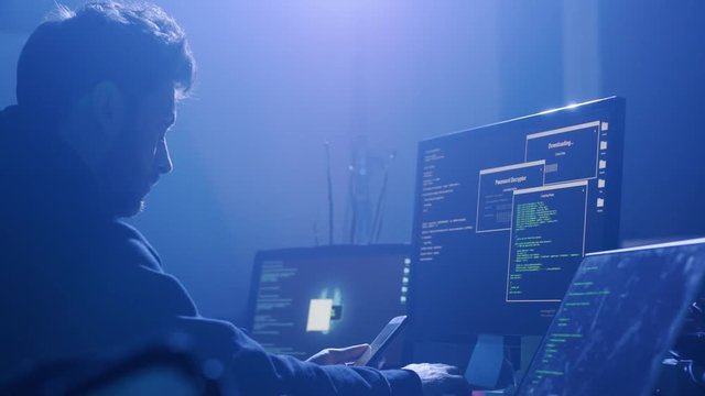 Profile of handsome smart computer hacker coding on computers at the desk in smoky room.