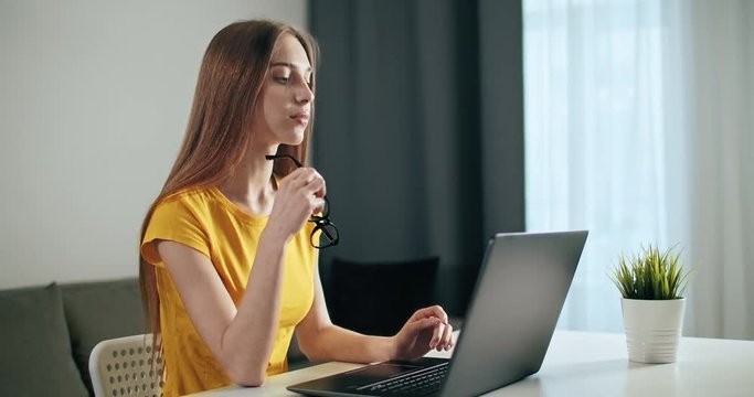 Young pretty woman dressed in yellow t-shirt and eyeglasses working on laptop, indoors shot in stylish apartment