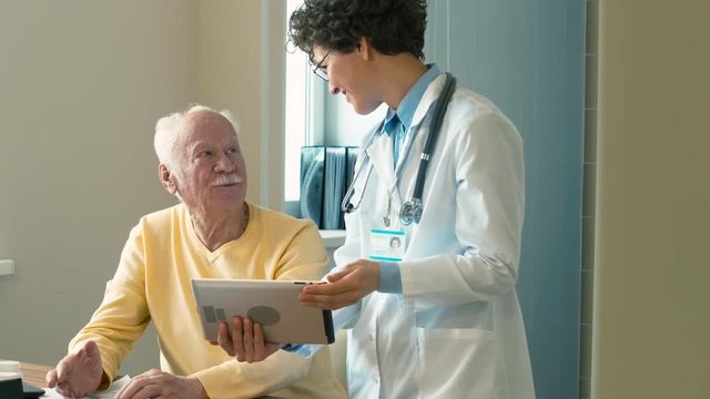 Doctor showing her elderly patient the results of analysis on tablet computer, and they glad because of successful treatment