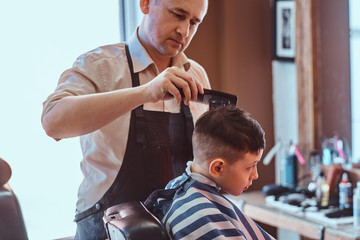 Young school boy is getting trendy hairstyle from expirienced hairdresser at modern barbershop.