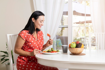 Beautiful brunette woman sitting wearing a red and white polka dot dress at dining table eating a healthy salad and drinking coffee or tea with a bowl of fruits on table with garden in the background