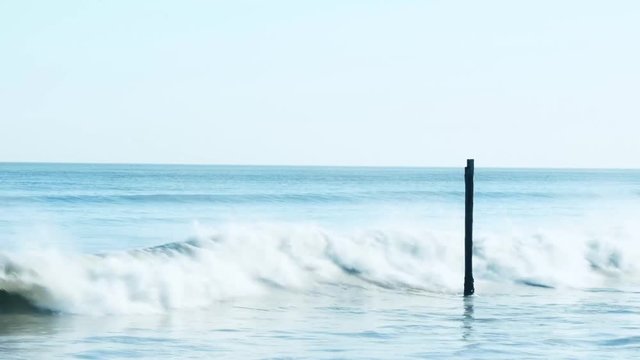 Migrating birds fly past an old pole in the ocean as it gets struck by the waves.