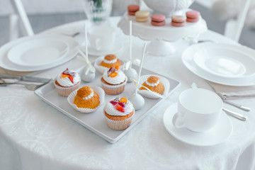 Creative spring composition. Elegant sweet dessert macarons, cup of tea or coffee and beautiful pastel colored beige and living coral flowers bouquet on white marble background
