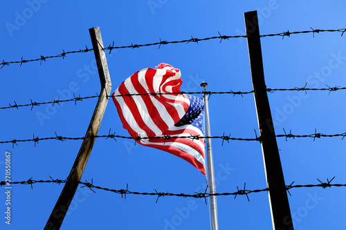 Conceptual image of waving American flag at tall pole and barbed wire fence over blue sunny sky