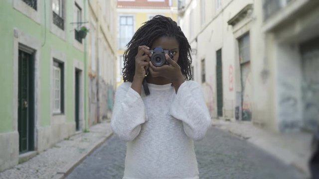 Smiling female photographer taking pictures on old city street. Beautiful African American woman with dreadlocks holding photo camera. Photography concept