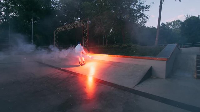 Aggressive Inline roller skater doing tricks in concrete skatepark with red burning signal flare outdoors, slow motion