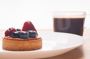 Closeup of delicious mini tart with berries and coffee cup. Beautiful pastry with strawberries and blueberries on plate on table. Breakfast or coffee break concept.
