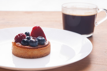 Closeup of delicious mini tart with berries and cup of coffee. Beautiful pastry with strawberries and blueberries on plate on table. Breakfast or coffee break concept.