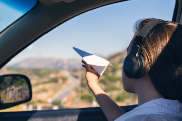 A girl in the headphones is launching a paper airplane from open window of the car during summer...