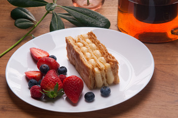 Closeup of napoleon cake portion and fresh berries on plate. Delicious pastry, teapot with tea and green leaves on wooden table. Breakfast or dessert concept.