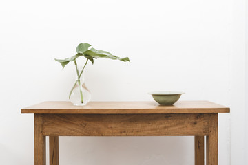 Monstera plant leaves in glass vase with green bowl on oak wooden table against white wall