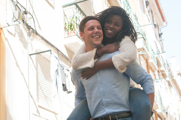 Smiling young man carrying girlfriend on back outdoors. Interracial couple in street. Romance and...