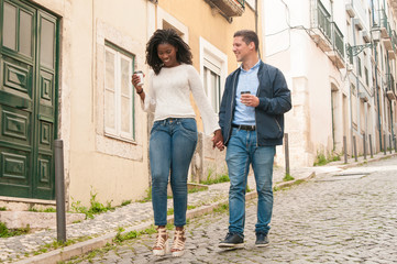 Happy Caucasian guy in love with her Afro American girlfriend. Mix raced couple holding hands, walking around old European city and drinking takeaway coffee. Dating concept - Powered by Adobe