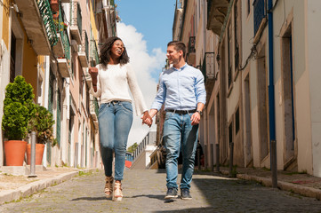 Fototapeta na wymiar Happy cheerful mix raced couple of tourists walking through old European city. African American girl and Caucasian guy holding hands, chatting and laughing. Multicultural relationship concept