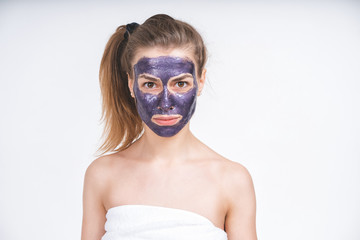 emotional portrait of a young beautiful european girl in violet cosmetic mask on her face. Wrapped in a towel. European, 22 years old, white, tail on the head, blank background, looking straight