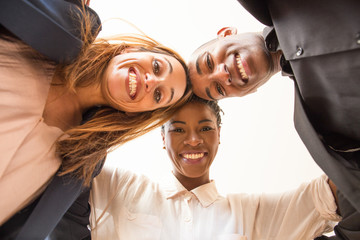 Low angle view of three embracing happy multiethnic colleagues. Portrait of African American businesswoman and businessman and Caucasian businesswoman smiling at camera. Business team concept