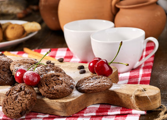 Oatmeal chocolate cookies with coffee grains and cherry, powdered sugar on kitchen cutting board with checkered fabric in village style for picnic. Macaron in background. It has pleasant taste.