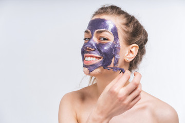 young girl with beautiful face removes a purple cosmetic mask. Close-up. Macro. Smile, joy. White beautiful teeth